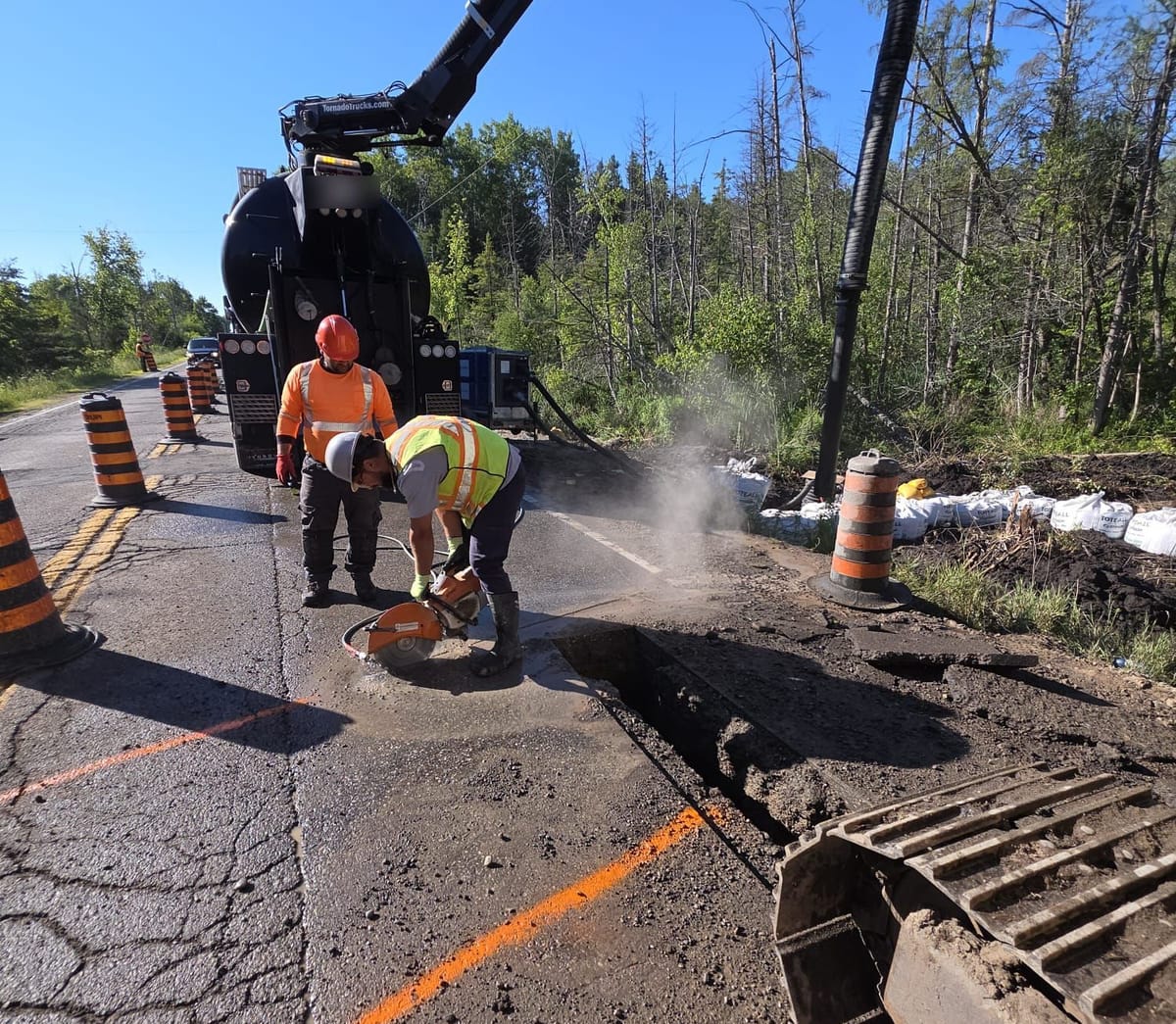 Two AMG crew in hard hats and hi-vis saw-cutting asphalt inside a coned lane before hydro excavation, with the truck boom overhead