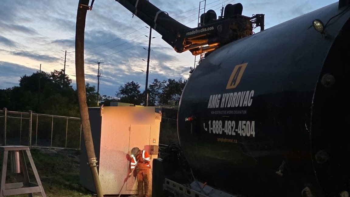 AMG Hydrovac truck at dusk with the boom extended to an operator excavating beside a rail-side equipment bungalow