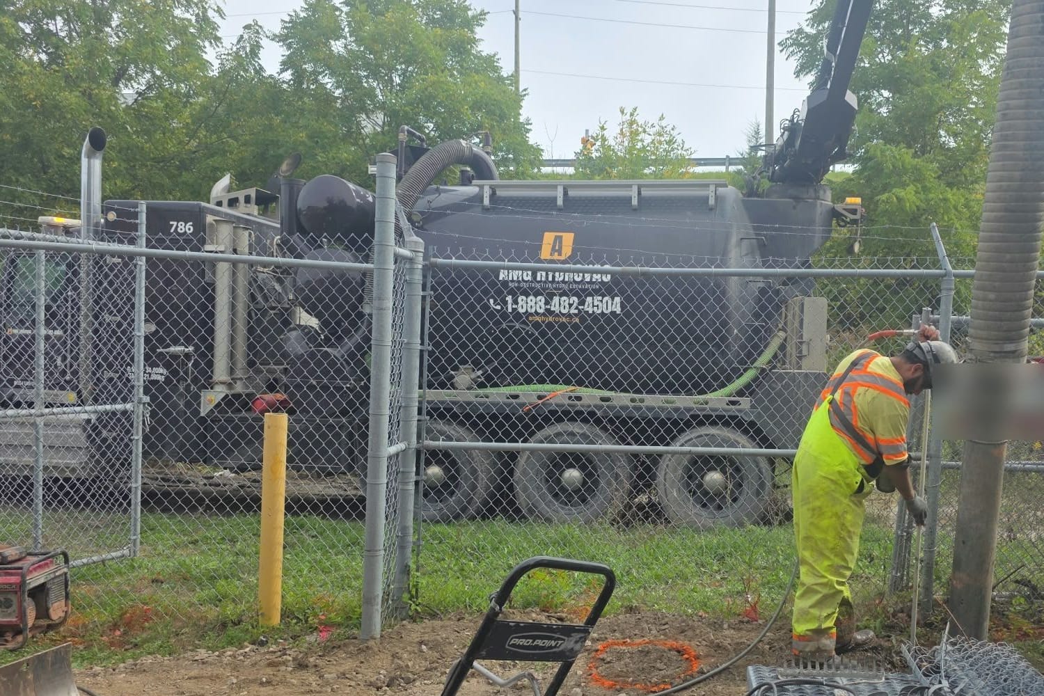 AMG Hydrovac truck parked outside a chain-link fence with the boom and hose reaching over to an operator digging inside the compound