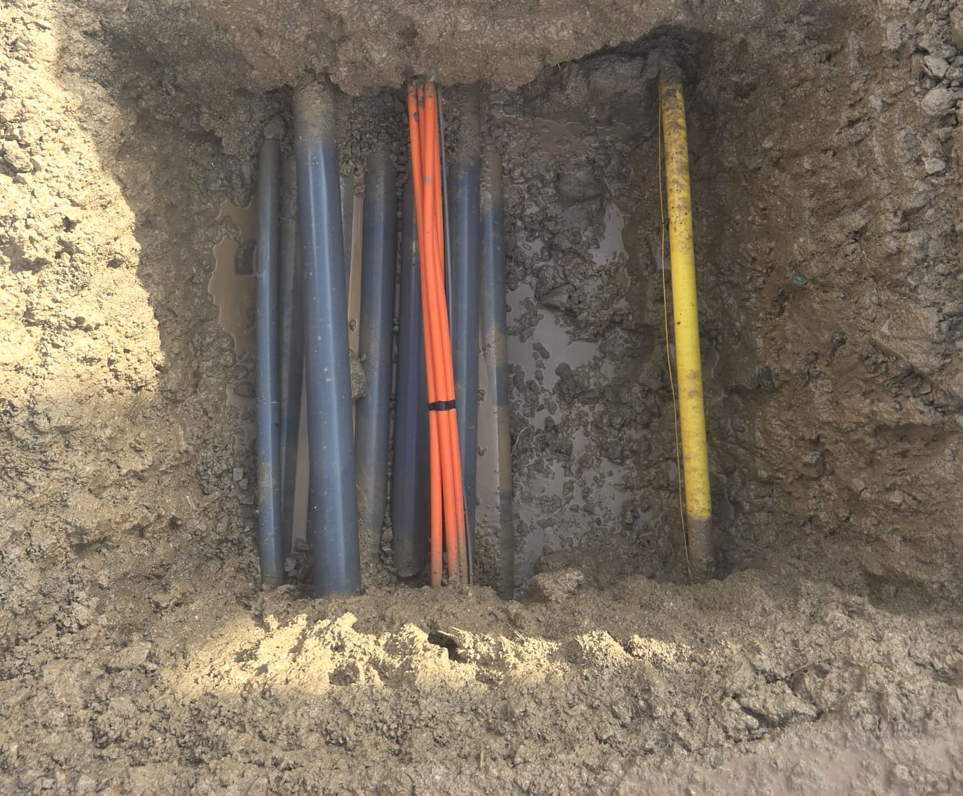 Square hydrovac test pit exposing a duct bank, a bundle of orange fibre conduits and a yellow gas line, all verified and intact