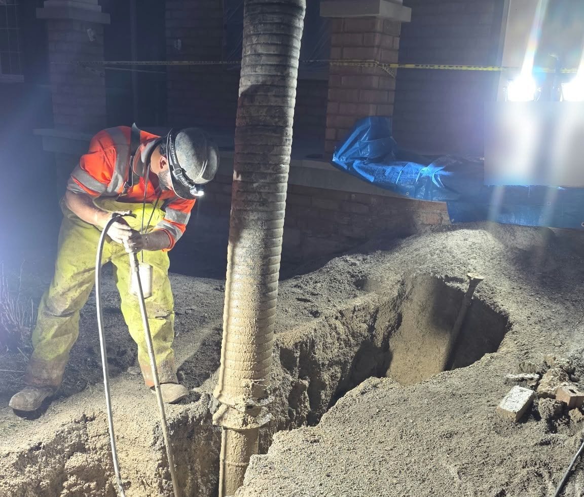 Operator with a headlamp working the hydrovac wand in an excavation at night under portable work lights
