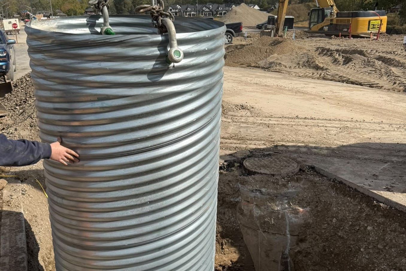 Corrugated steel pipe sleeve being lowered by excavator chains into a hydrovac-dug pole hole while the crew guides it