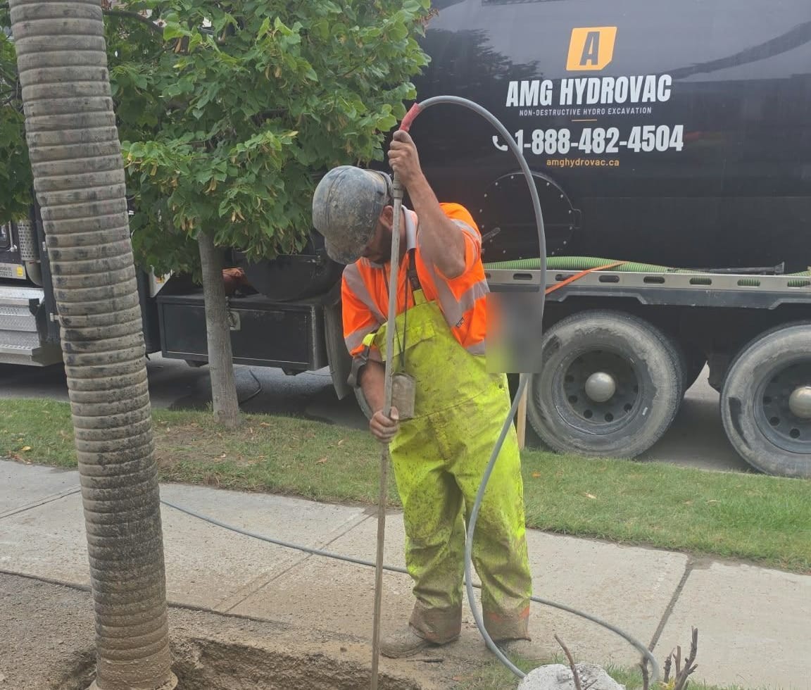 AMG operator in a hard hat and hi-vis working the hydrovac wand in a boulevard pothole, the AMG Hydrovac truck parked at the curb behind him