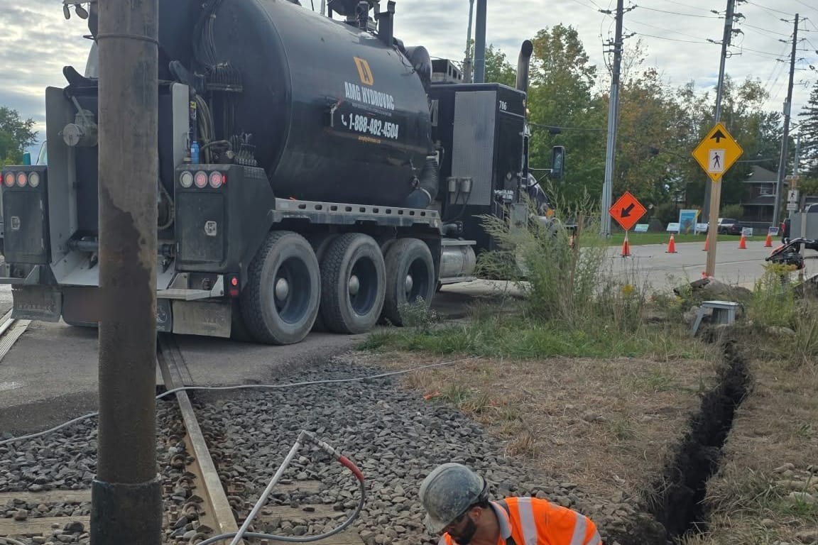 AMG crew member in hi-vis excavating a narrow trench across railway ties with the AMG Hydrovac truck parked at the crossing