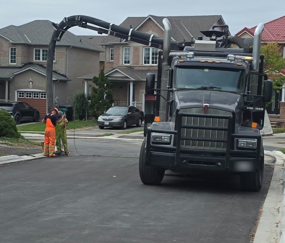 AMG Hydrovac truck parked on a residential street with its boom extended to a crew excavating at the curb