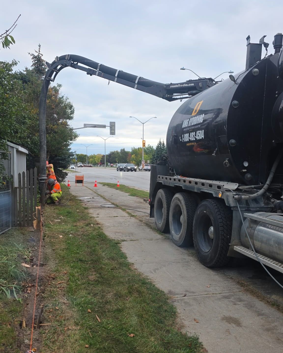 Side view of an AMG Hydrovac truck with its boom extended over the sidewalk to a crew digging along a fence