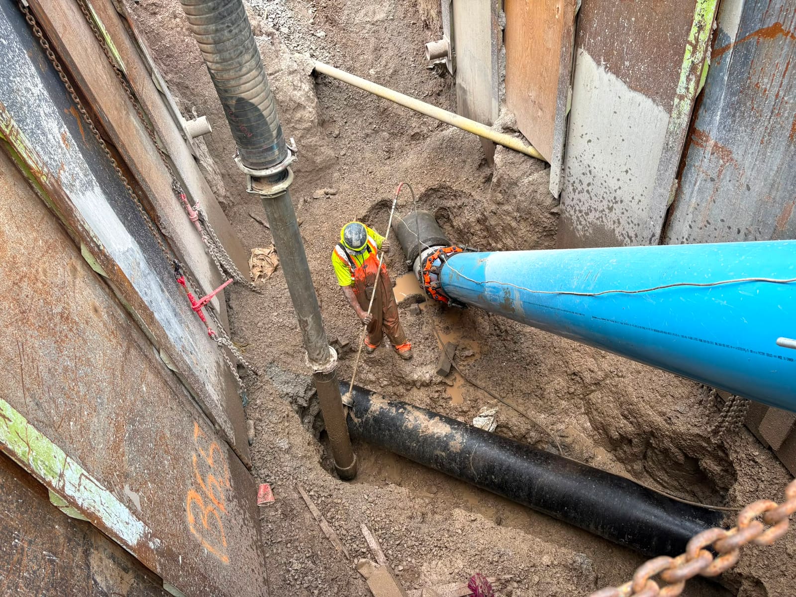 AMG operator in full PPE hand-wanding spoil away from a live watermain tie-in at the base of a fully shored excavation